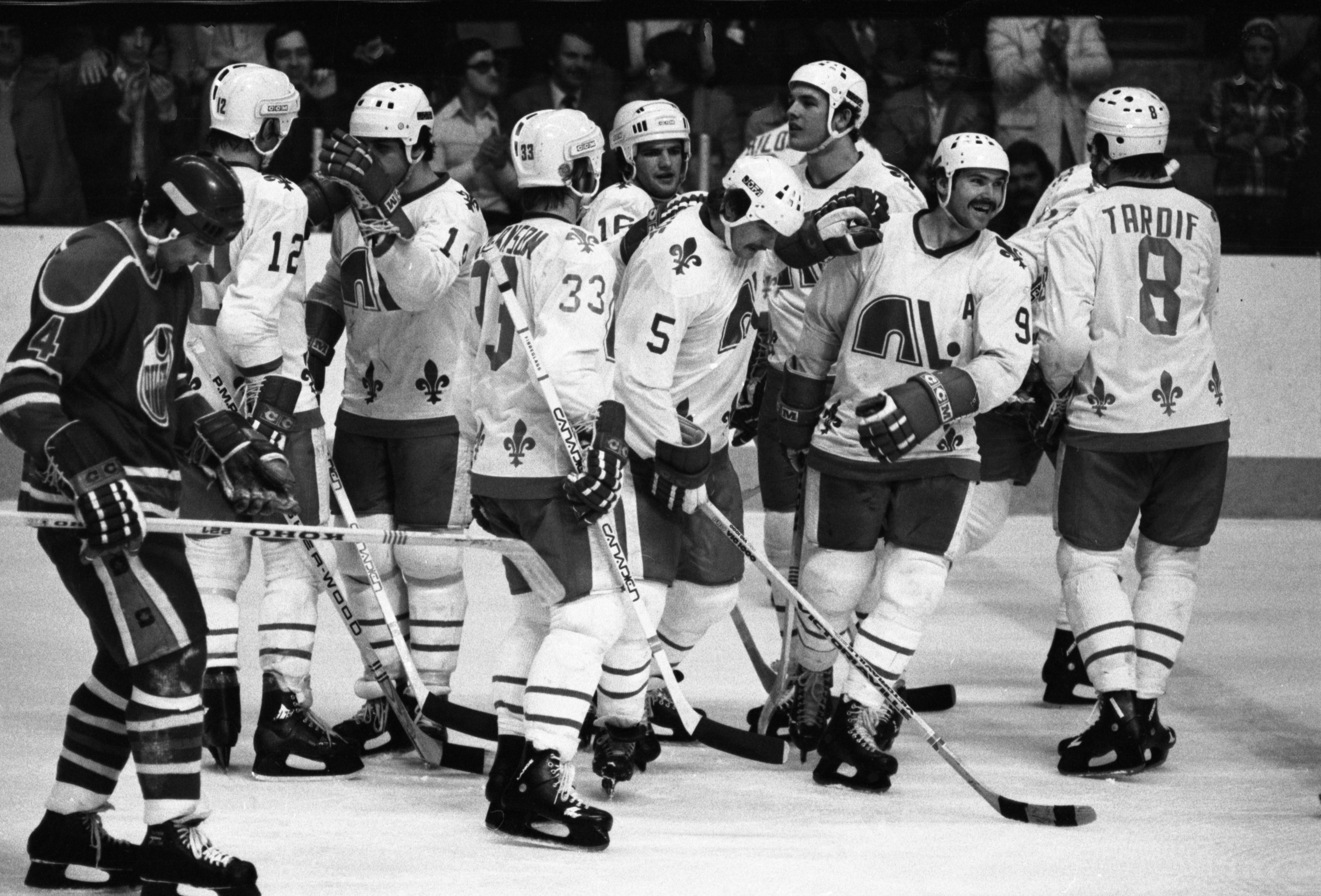 Photographie présentant neuf joueurs des Nordiques et un joueur des Oilers en équipement de hockey sur la patinoire. Les Nordiques de Québec célèbrent un but marqué contre les Oilers d’Edmonton au Colisée de Québec. Les deux équipes font le saut dans la Ligue nationale de hockey (LNH) lors de la dissolution de l’AMH en 1979. Parmi les joueurs des Nordiques figurent Réal Cloutier, Marc Tardif et Dale Hoganson. Cloutier et Tardif sont parmi les meilleurs joueurs de la ligue.
