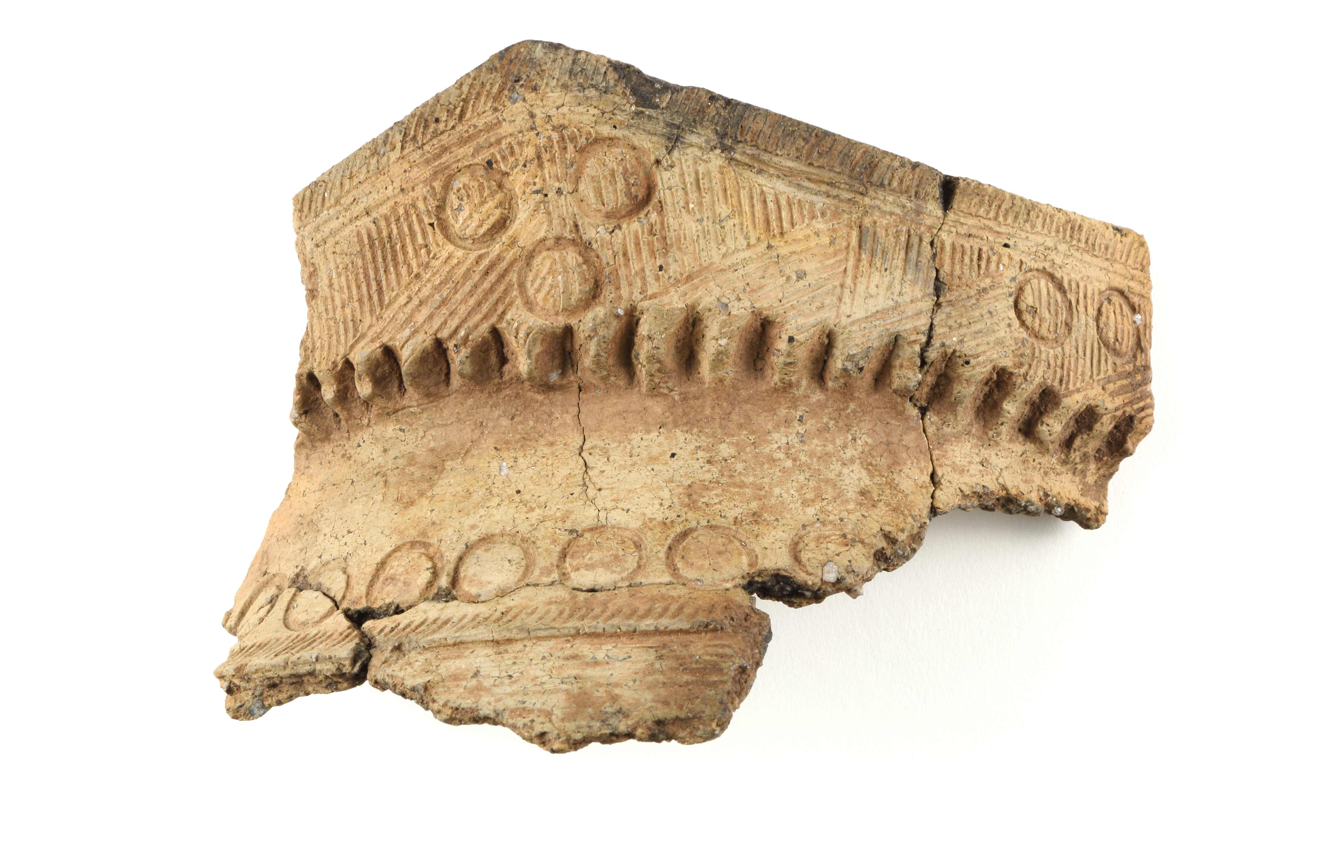 Photographie sur fond blanc d’un fragment de vase d’argile décoré de motifs gravés. Le fragment mesure environ cinq centimètres. Les archéologues retrouvent plusieurs fragments de vases provenant de la communauté iroquoienne locale sur le site de Charlesbourg-Royal. Les décorations du vase suggèrent qu’il provient de Stadaconé. Il s’agit de la localité iroquoienne voisine de la colonie française. Le positionnement des fragments dans les couches de sol indique qu’ils datent de l’occupation française, ou peu de temps après.
